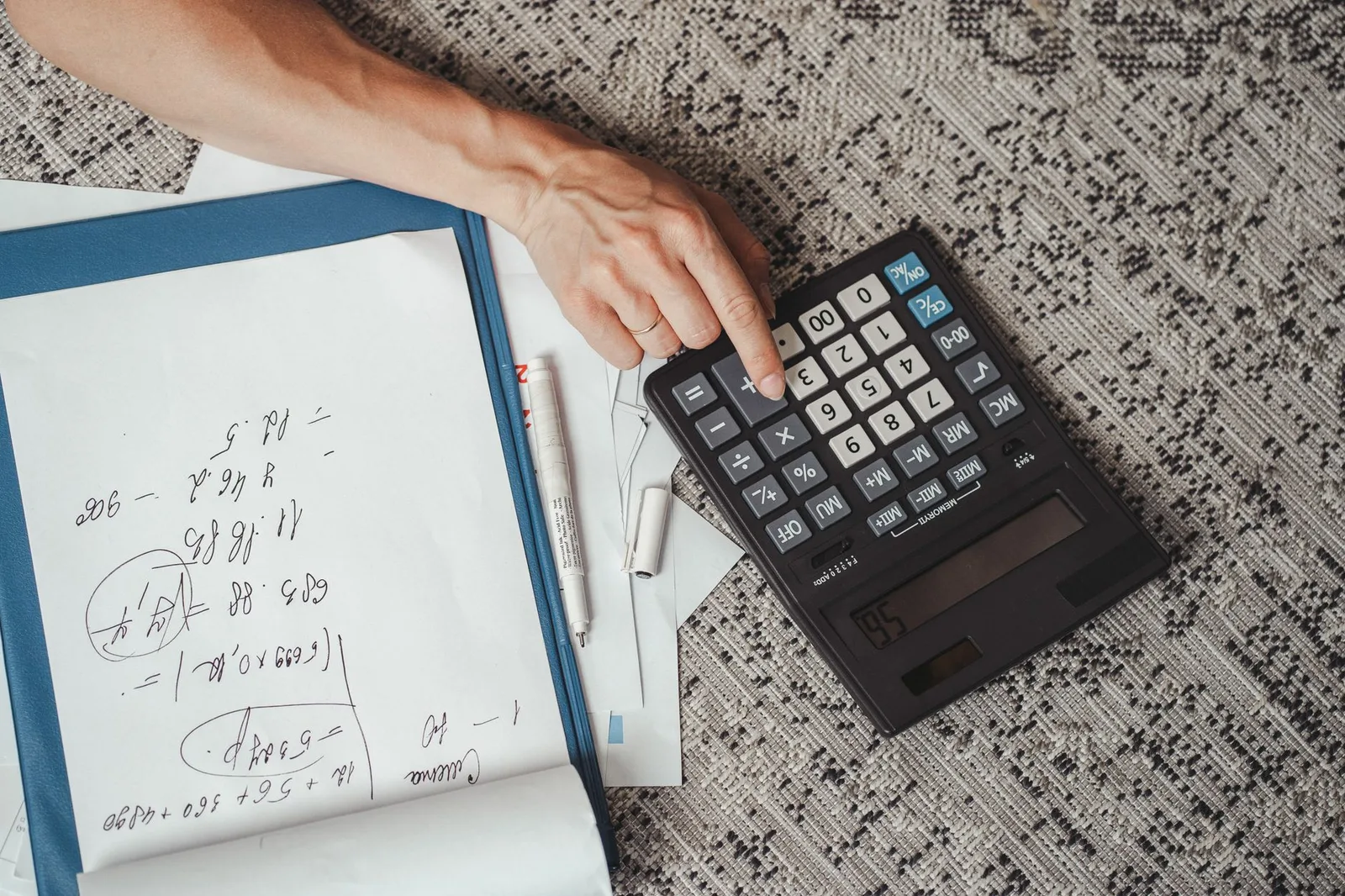 A hand using a large calculator beside handwritten notes and papers on a desk, seen from above.