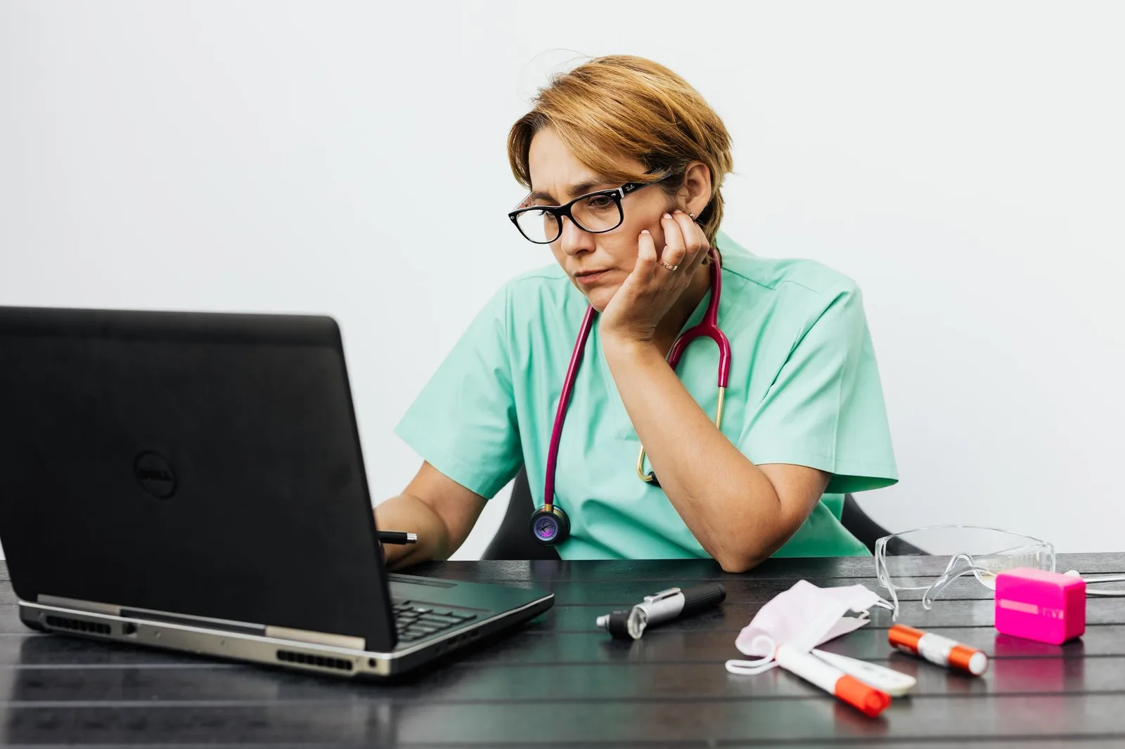 A nurse wearing glasses working at a laptop on a desk, with medical instruments beside it.