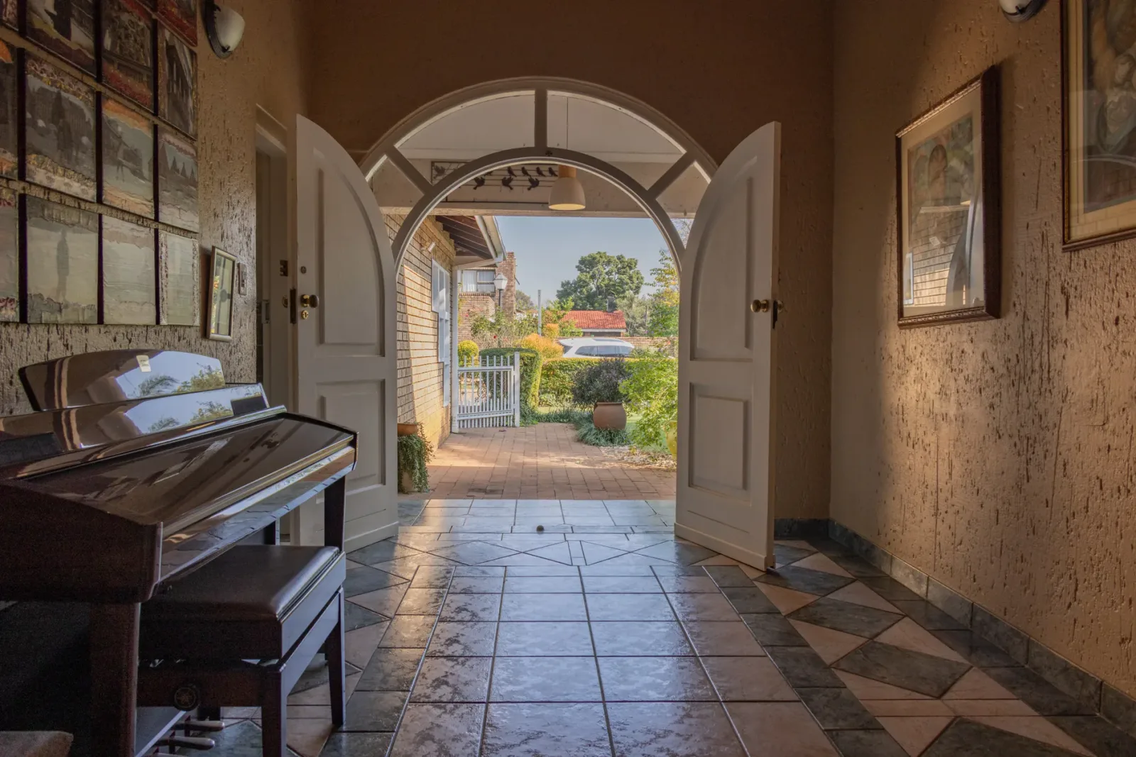 A tiled hallway with tall doors standing open onto a sunlit garden, an upright piano against the wall.