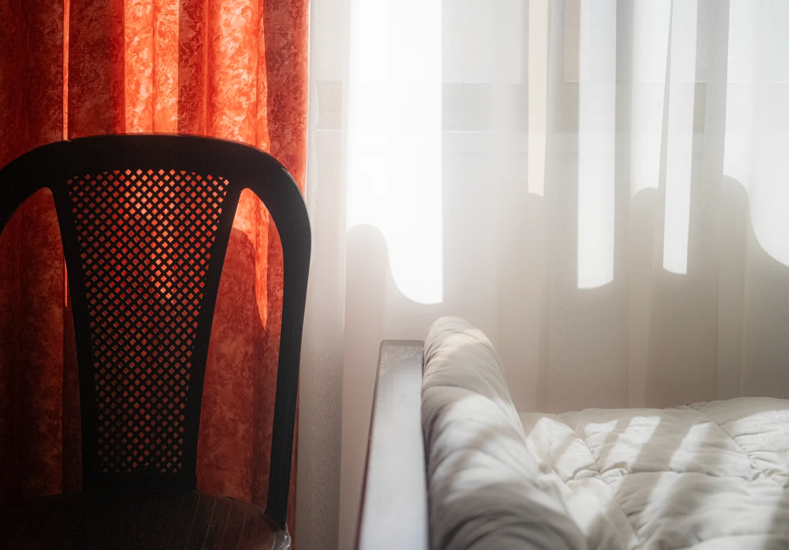 Sunlight through a sheer curtain falls across an empty single bed and a chair in a quiet room.