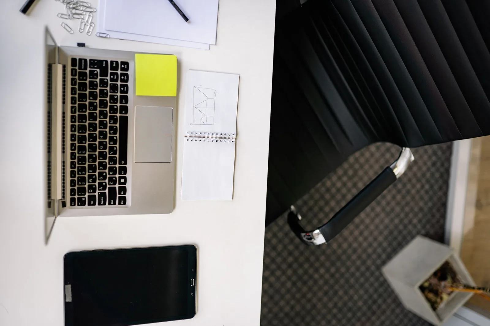 A tidy office desk seen from above, with an open laptop, a tablet and a notepad.