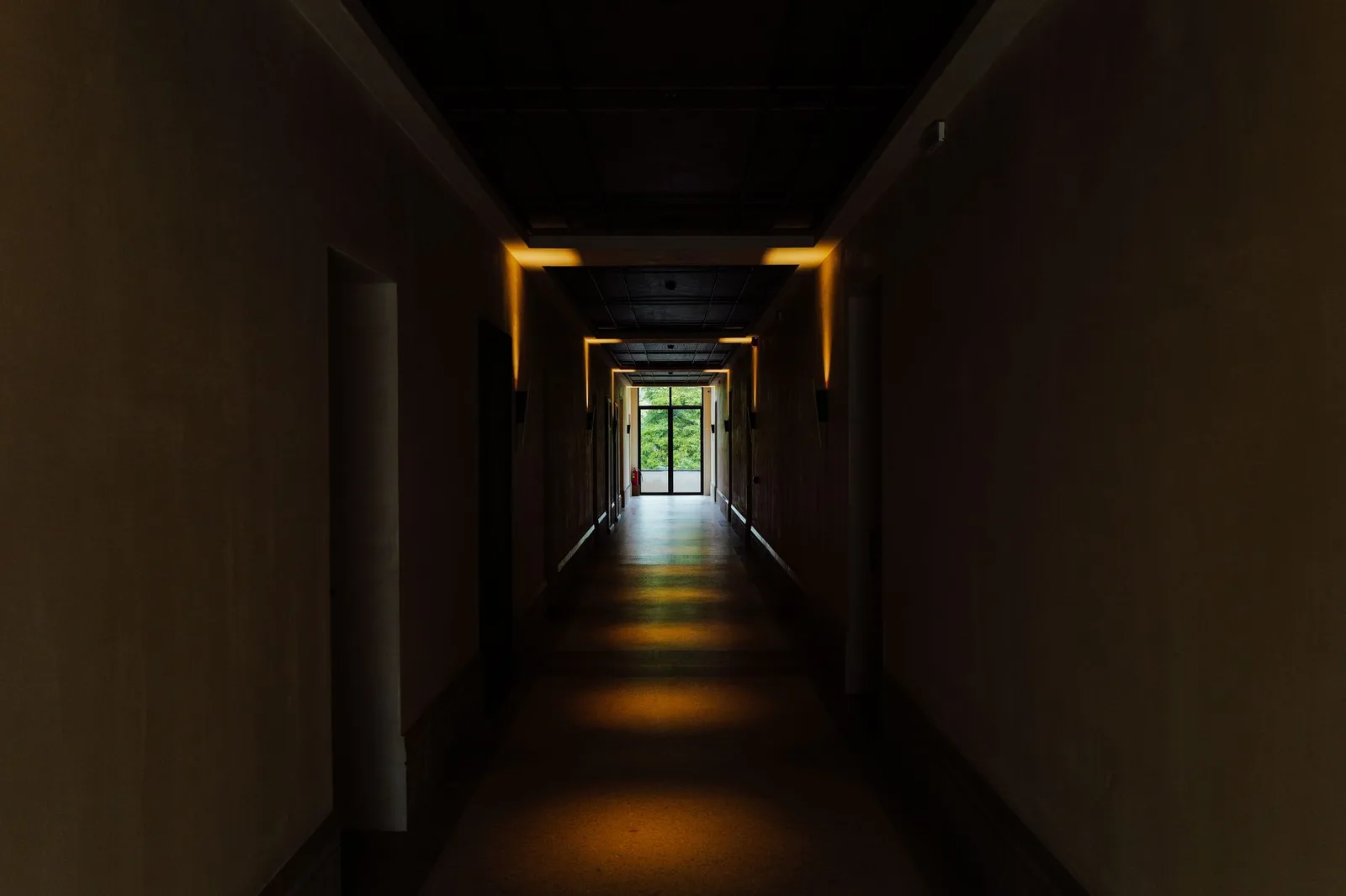 A dim, empty indoor corridor lit by low wall lights, with daylight showing through a doorway at the far end.