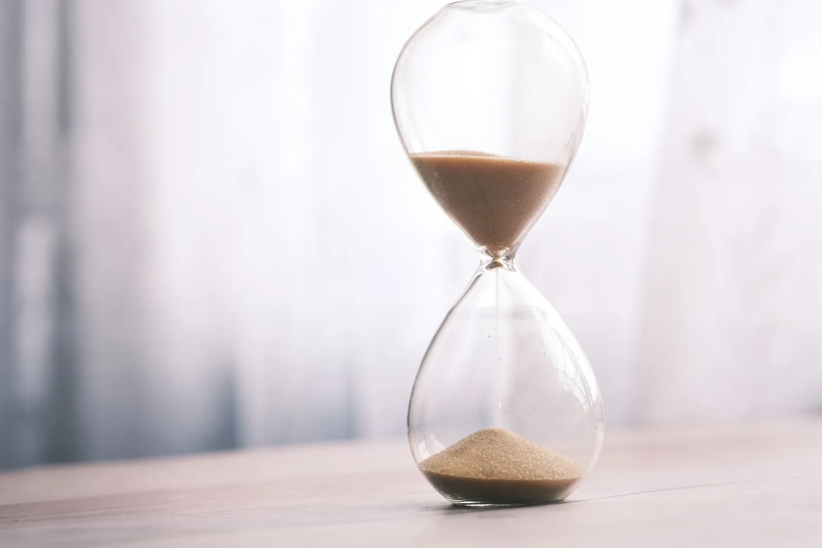 An hourglass in soft daylight, sand part way through, with pale light behind it.